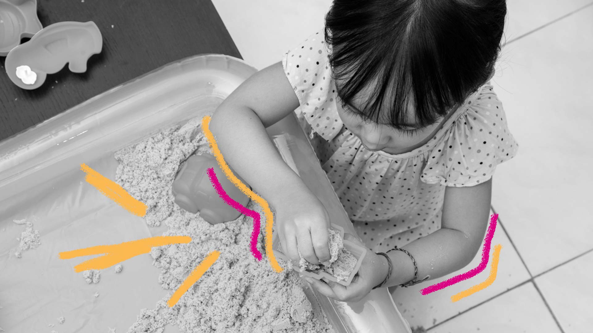 Areia caseira: foto preto e branco de uma criança, que está brincando com uma caixa de areia. Há rabiscos em rosa e laranja na imagem.