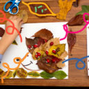 Em primeiro plano, a foto de uma mesa vista de cima, por onde estão espalhadas folhas de diferentes cores e tamanhos que, juntas, formam o desenho de um animal.. A mão de uma criança segura uma das folhas.
