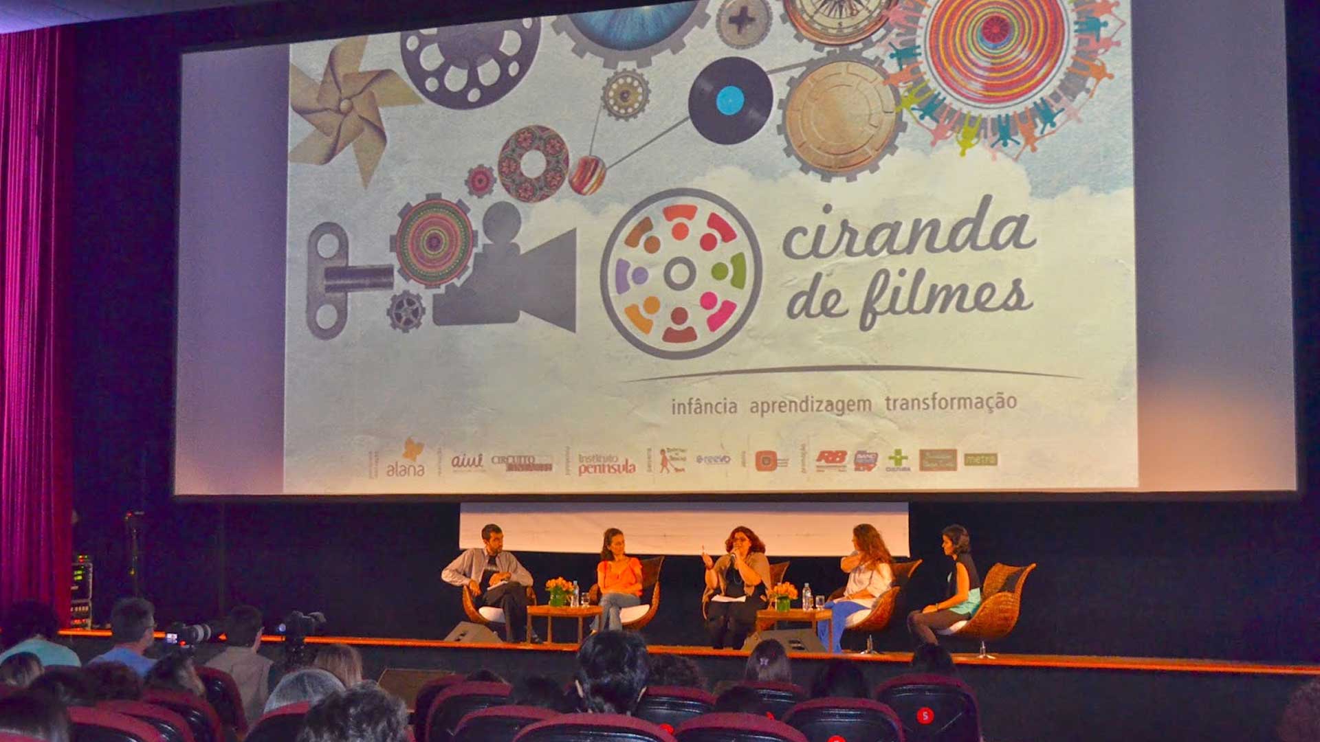 Foto de um palco de uma sala de cinema, cinco pessoas discursam. Acima, um telão com o logo do projeto Ciranda de Filmes.