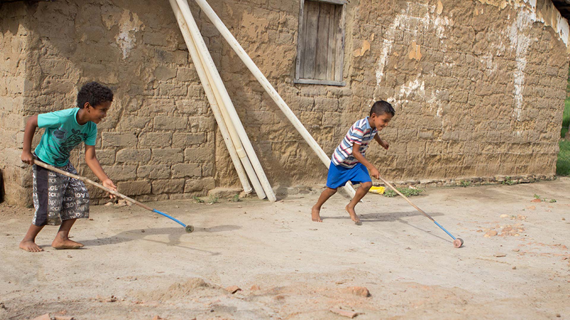Dois meninos pequenos brincando descalços com pedaços de madeira em frente a uma casa simples e chão de terra batida
