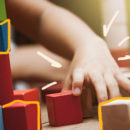 Em primeiro plano, as mãozinhas de uma criança brincando com blocos coloridos de madeira.