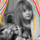 Duas meninas brincam debaixo de uma cabaninha de pano montada na sala. Grafismos coloridos enfeitam a imagem.