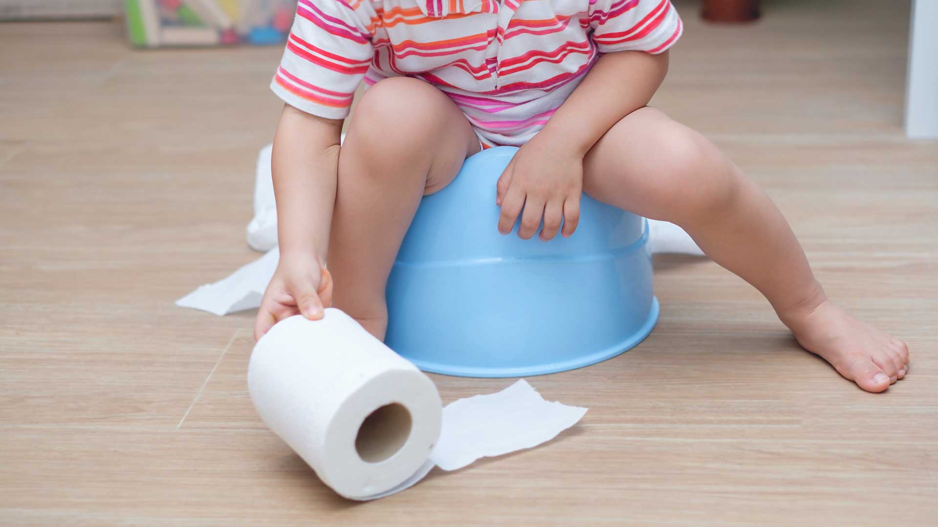 Uma criança, do tronco para baixo, sentada em um penico azul, esticando a mão para alcançar um rolo de papel higiênico
