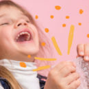 Criança sorri apertando um pedaço de plástico bolha, sob um fundo cor-de-rosa.
