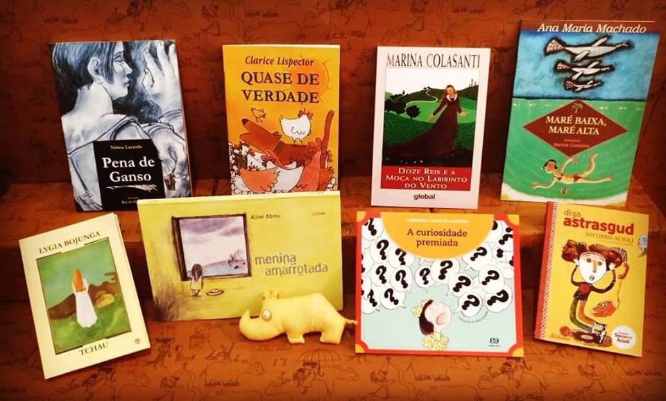 Livros infantis expostos em pé em uma mesa, ao lado de um pequeno rinoceronte de plástico, mascote do Clube Quindim