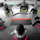 Foto em preto e branco. Em uma sala, várias pessoas reunidas em círculo sentadas em bolas de yoga.