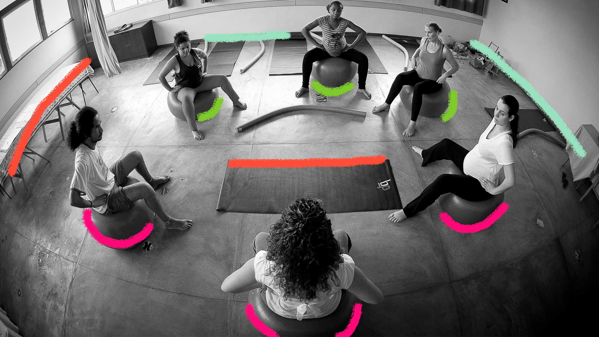 Foto em preto e branco. Em uma sala, várias pessoas reunidas em círculo sentadas em bolas de yoga.