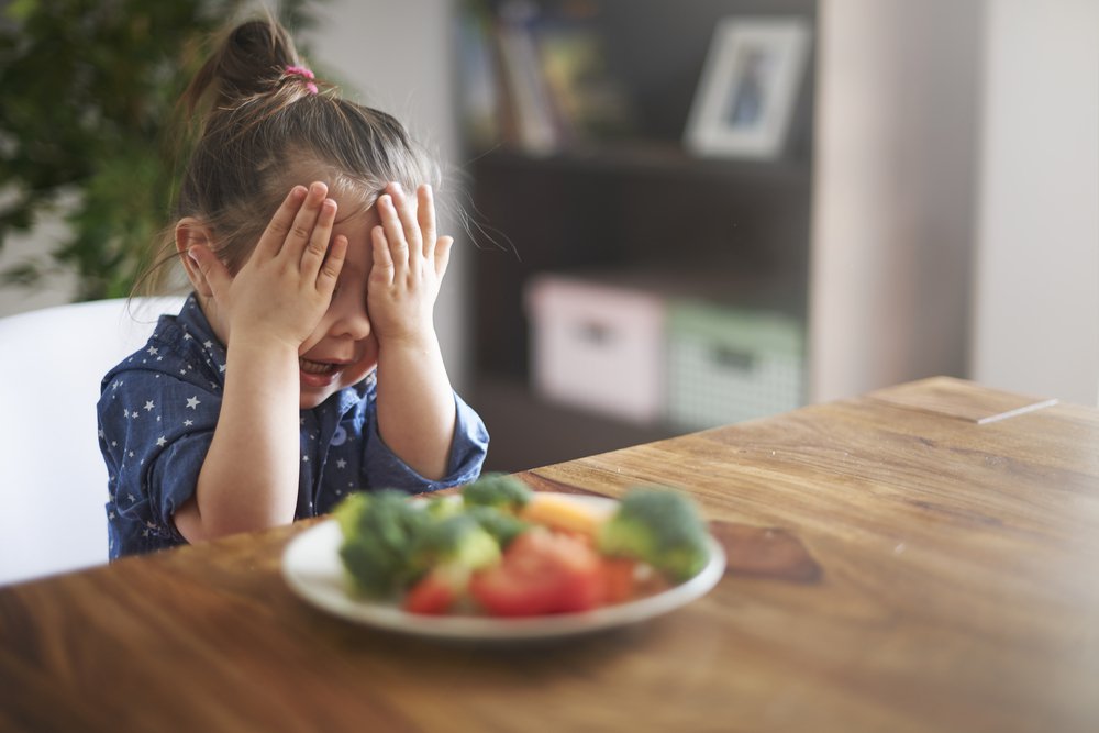 Criança sentada à mesa diante de um prato de legumes. Ela está tapando os olhos com as mãos e chorando.