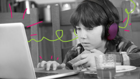 TDAH: Menino em frente ao notebook olha concentrado a tela e utiliza um headfone roxo nos ouvidos.