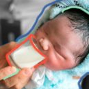 um bebê recém-nascido de olhos fechado segurado por um adulto oferecendo um pequeno copo de leite