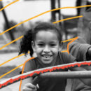 Rosto de menina sorrindo, com o cabelo preso em rabo de cavalo, brincando no gira-gira de uma escola. Foto em preto e branco.