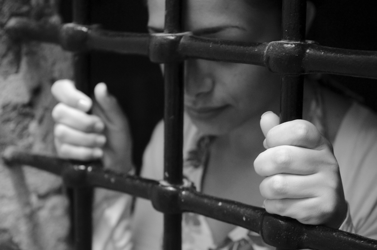 Mulher na prisão segura as grades com as duas mãos e encosta a testa sobre o ferro.