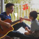 Músicas de pai para filho: na foto, pai e filha de etnia amarela se olham. O pai toca violão, usa óculos e camiseta azul, enquanto a filha segura uma caneca na mão.