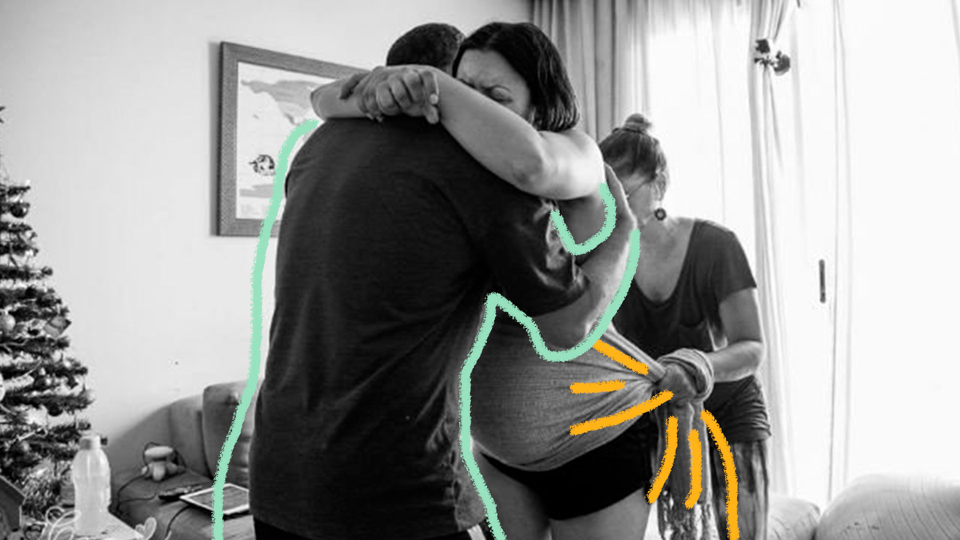 Uma foto em preto e branco de uma mulher grávida abraçada a um homem. Atrás deles, uma mulher amarra um xale de tecido em sua barriga.