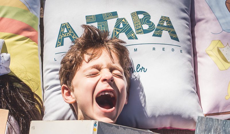 Um menino está deitado com um livro na mão, em um travesseiro onde está escrito "A Taba"