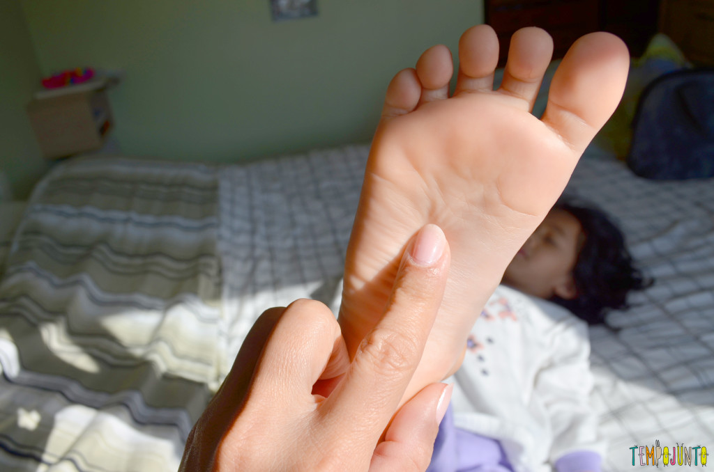 Criança deitada na cama com o pé para cima. Um adulto passa os dedos pela sola do pé da criança.