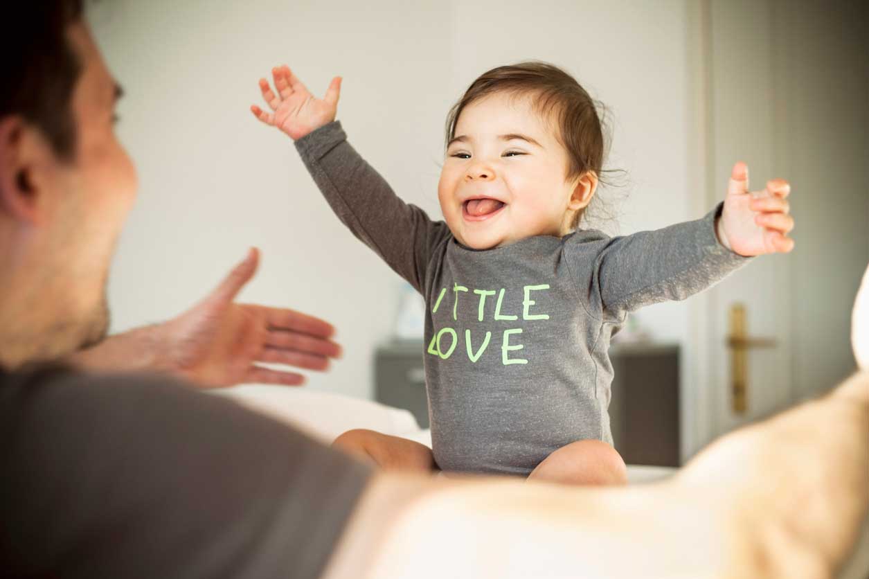 homem adulto brinca com bebê no colo, com os braços abertos e sorrindo.