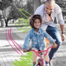 Em um parque, um garoto sorri andando de bicicleta enquanto seu pai o incentiva logo atrás.