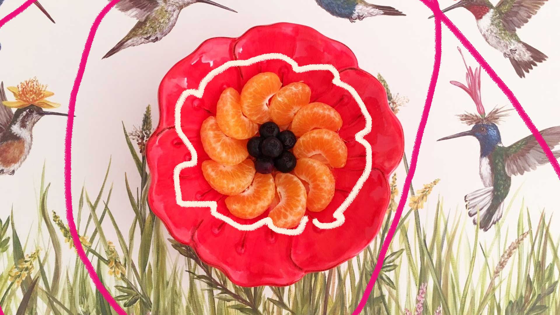 Em uma mesa com uma toalha de beija-flores, no centro uma travessa vermelha contém gomos de laranja dispostos como uma flor.
