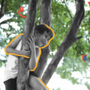 Menino abraça um tronco no alto de uma árvore e sorri. Ao fundo da foto podem ser vistas as folhas verdes de outras árvores, o que nos remete ao contato com a natureza.