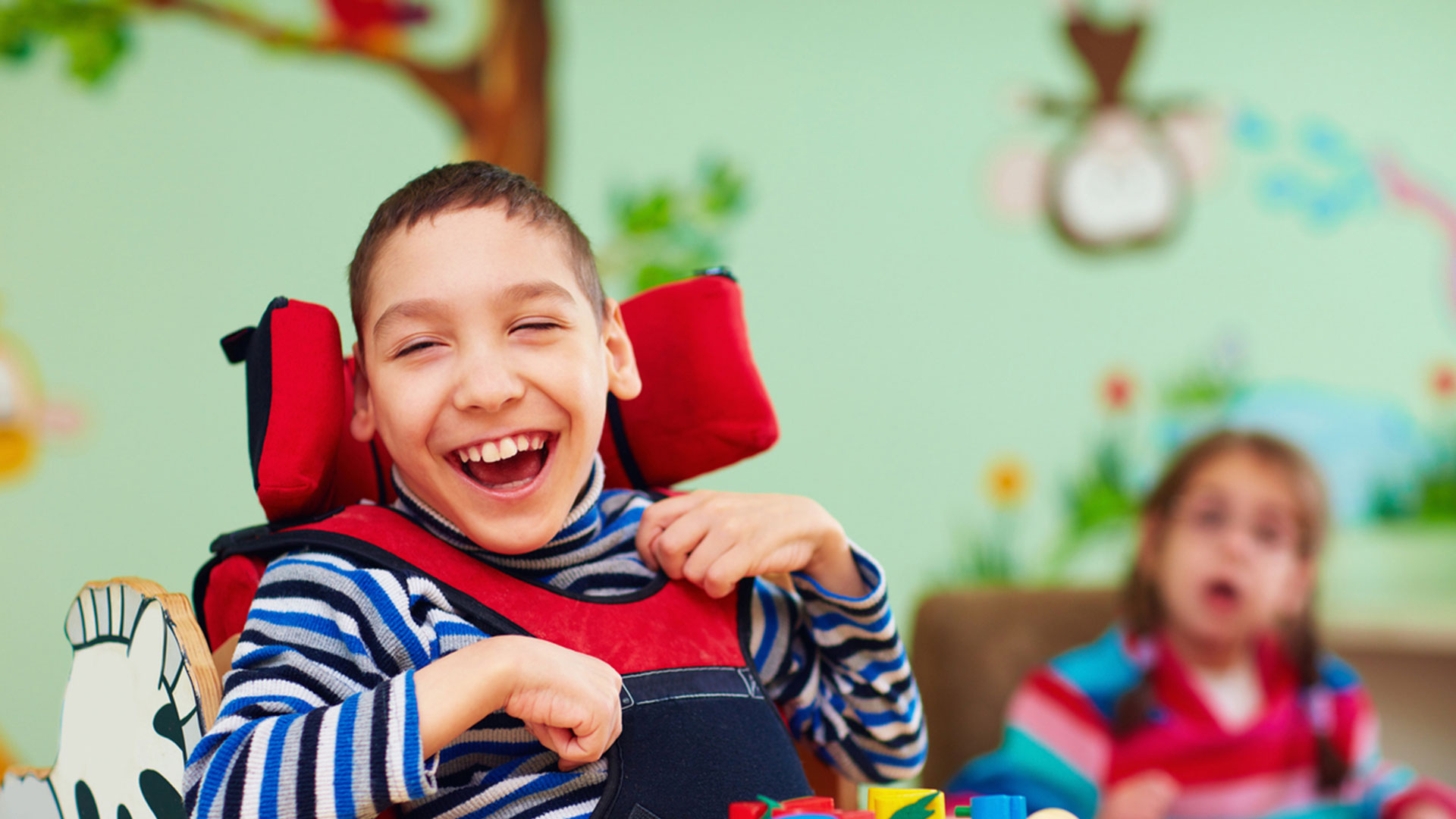 Menino com deficiência sentado em cadeira de rodas sorri largamente em uma sala de brincar colorida