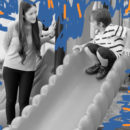 Foto em preto e branco mostra uma professora em pé olhando um menino, que está brincando no escorregador.