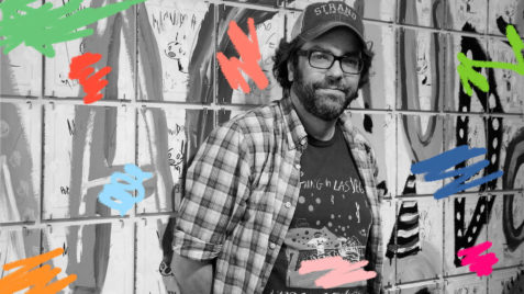 Foto em preto e branco mostra o artista Liniers vestindo uma camisa xadrez aberta, com camiseta por baixo, óculos de grau e um boné.