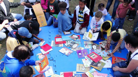 Crianças se agacham no chão em uma toalha estendida, com diversos livros espalhados por cima.