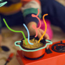 Mãos de uma criança brincando com um fogãozinho. Em cima de uma das bocas do fogão, existe uma panela com macarrão e água dentro.