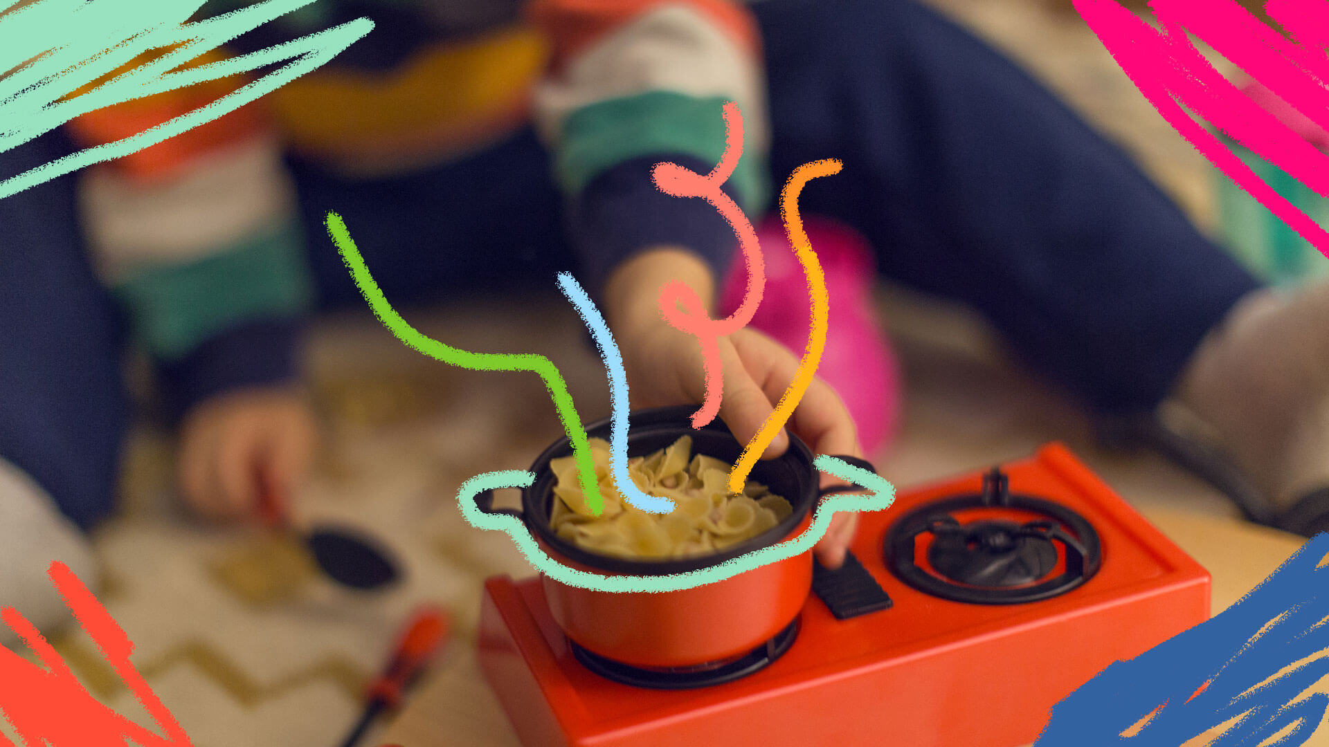 Mãos de uma criança brincando com um fogãozinho. Em cima de uma das bocas do fogão, existe uma panela com macarrão e água dentro.