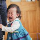 Criança asiática chorando e se agarrando à perna de um adulto.