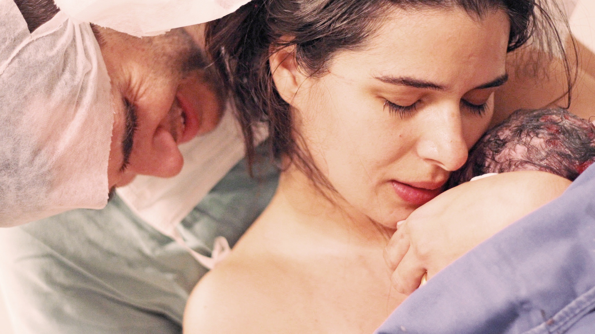 Mãe acaba de dar à luz, com bebê no colo. Ao seu lado, um homem com touca hospitalar lhe abraça e chora.