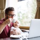 Homem de óculos sentado em uma mesa em frente a umcomputador segurando uma xícara de café com um bebê no colo