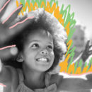 Foto em preto e branco mostra o rosto de uma menina sorrindo, com as duas mãos para o alto.
