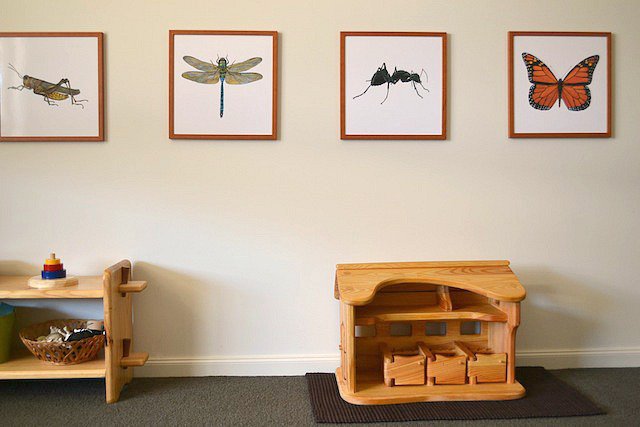Quarto montessoriano infantil: foto de uma parede, que tem quadros pendurados e uma caixa de madeira apoiada ao chão.