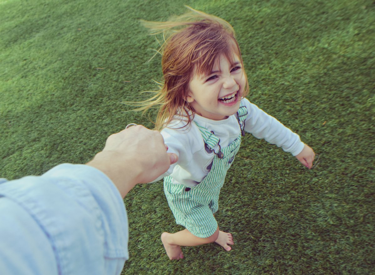 Uma menina loirinha está sorrindo e correndo em um gramado, segurando a mão de sua mãe (só vemos o braço do adulto), enquanto é fotografada de cima