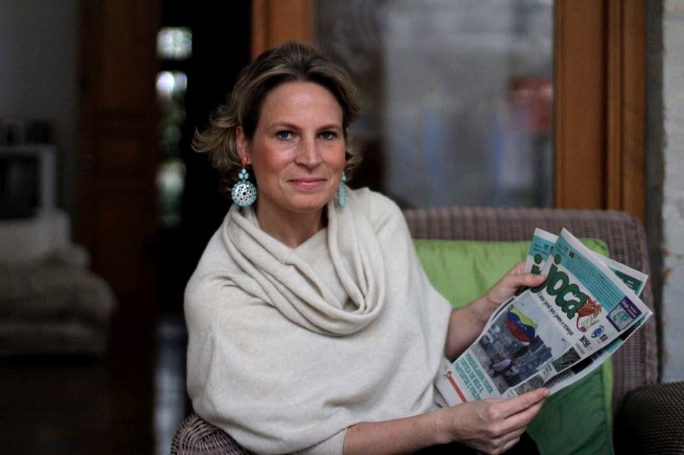 Foto de uma mulher loira, de pele clara, vestindo uma blusa branca e sentado em um sofá com uma edição do jornal O Joca nas mãos