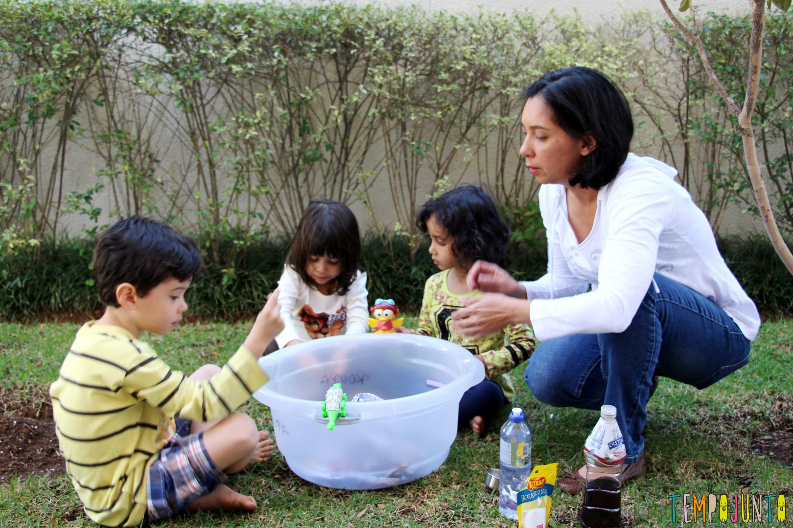 Como fazer um vulcão em casa: foto de três crianças e uma mulher adulta, que estão em um quintal e mexem em uma bacia.