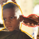 Rosto de menino negro com o cabelo curto sendo cortado por uma máquina, Aparece somente a mão de quem corta.