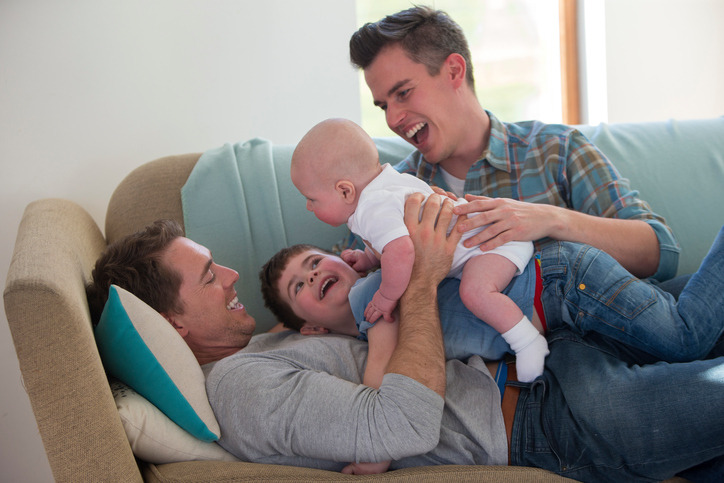 Foto de um casal homoafetivo que brinca com os dois filhos no sofá.