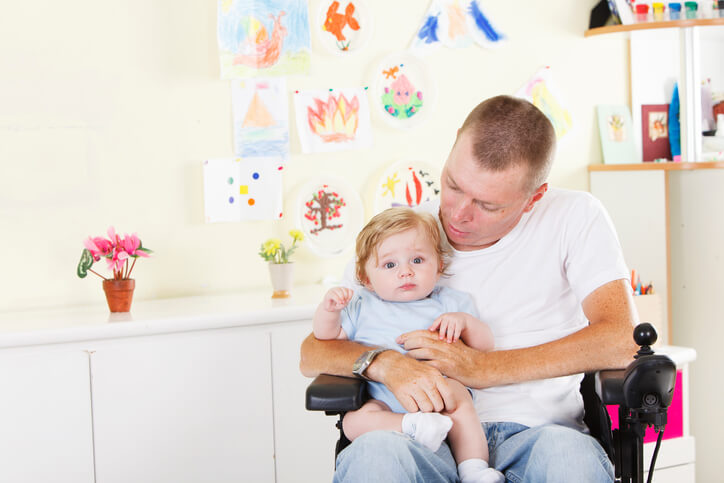 Foto de um homem cadeirante segurando um bebê no colo. Na parede, atrás, há desenhos feito por crianças