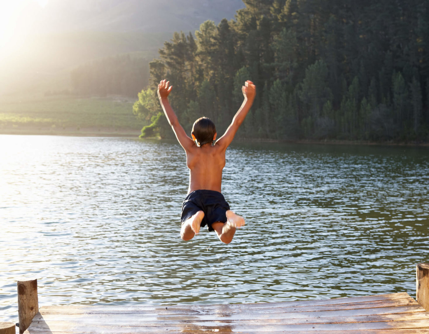 Um menino está saltando em um rio de braços abertos. O dia está ensolarado e o cenário é de mata fechada