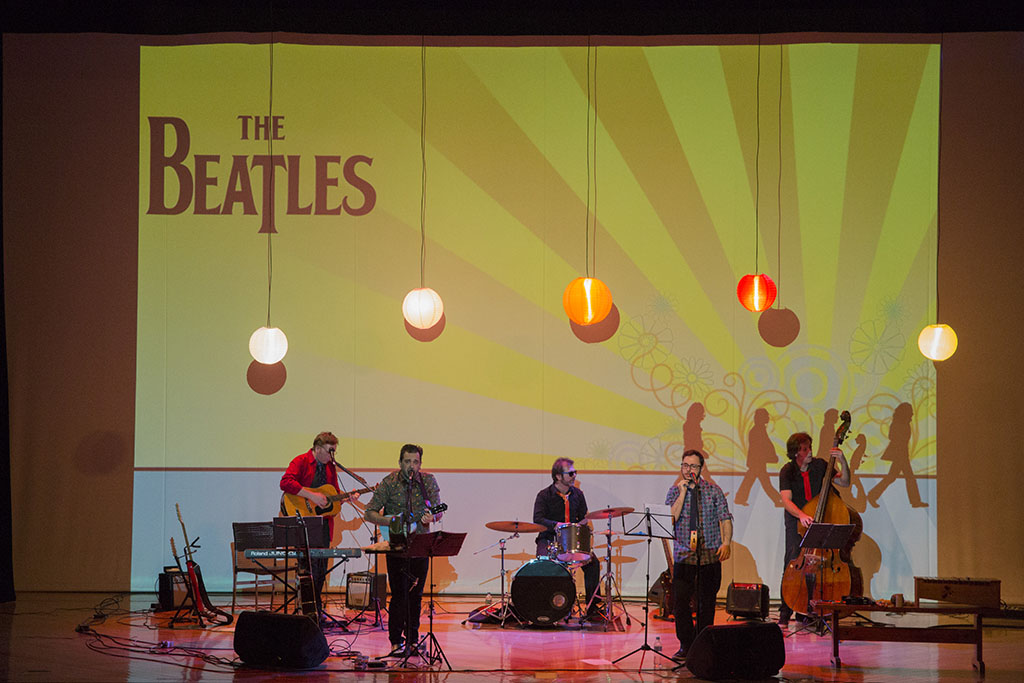 A banda cover Beatles para Crianças em um palco iluminado por luzes coloridos e com uma projeção dos Beatles ao fundo.