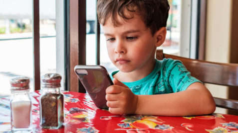 Menino de pele branca segurando e olhando para um celular em sentado em uma mesa. Vê-se potes de vidro de tempero