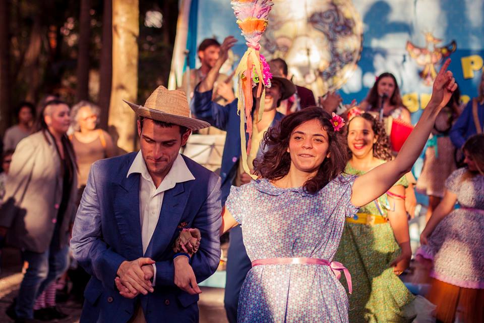 Um casal de mãos dadas vestido à caráter com trajes juninos dançam em uma quadrilha