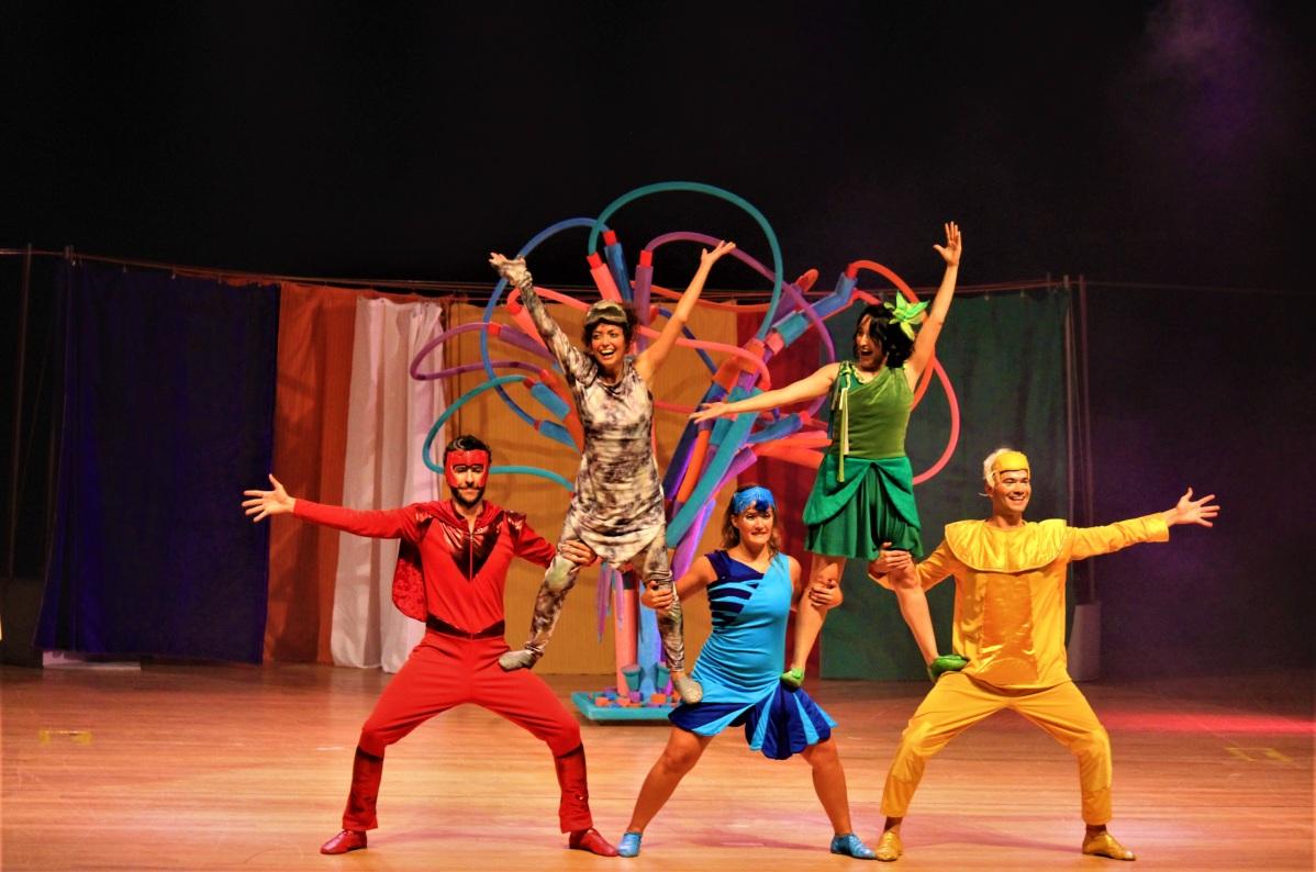 Em um palco, cinto atores vestidos cada um de uma cor do arco-íris fazem uma pirâmide humana, com os braços estendidos e sorrindo.
