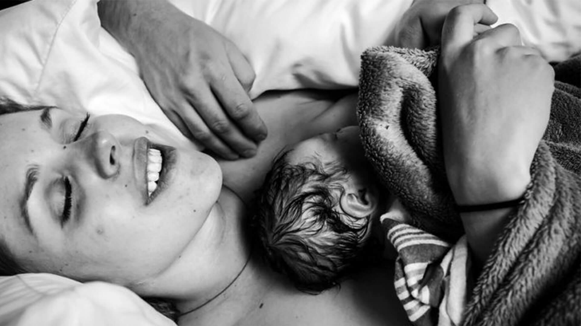 Foto em preto e branco mostra mãe e bebê deitados. O recém-nascido está no colo da mãe e ela sorri.