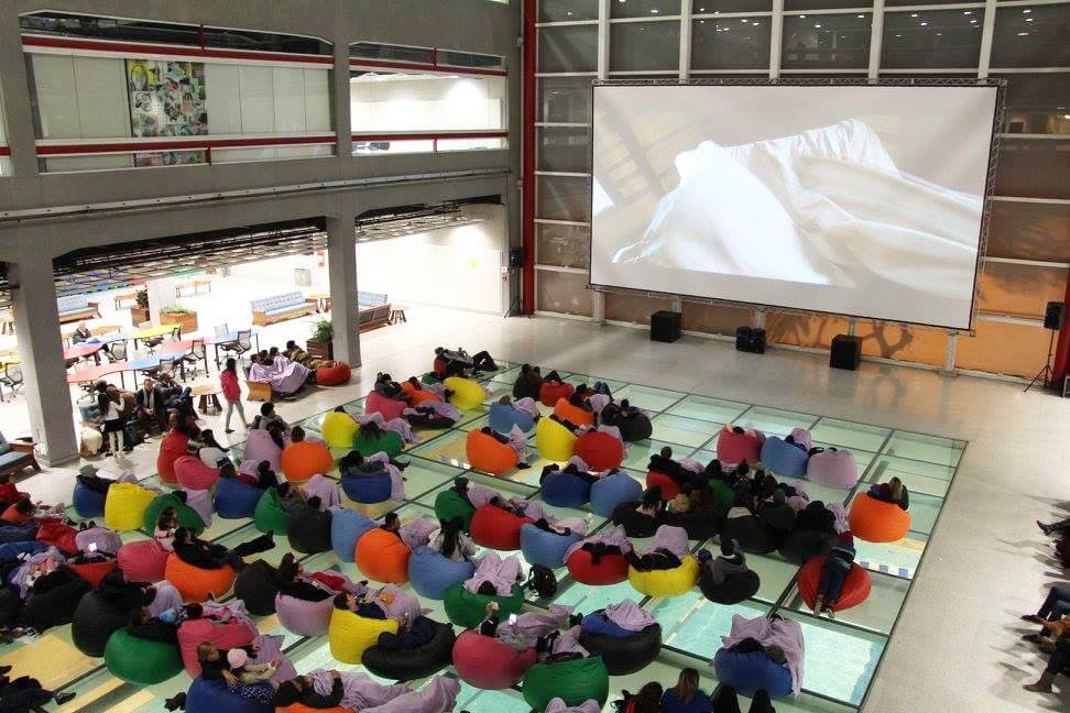 Pessoas sentadas em pufes coloridos em cima do chão de vidro olhando para a tela do cinema