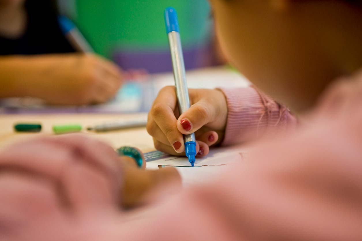 Criança com roupa cor-de-rosa escrevendo com caneta azul e branca. Foto tirada em close.