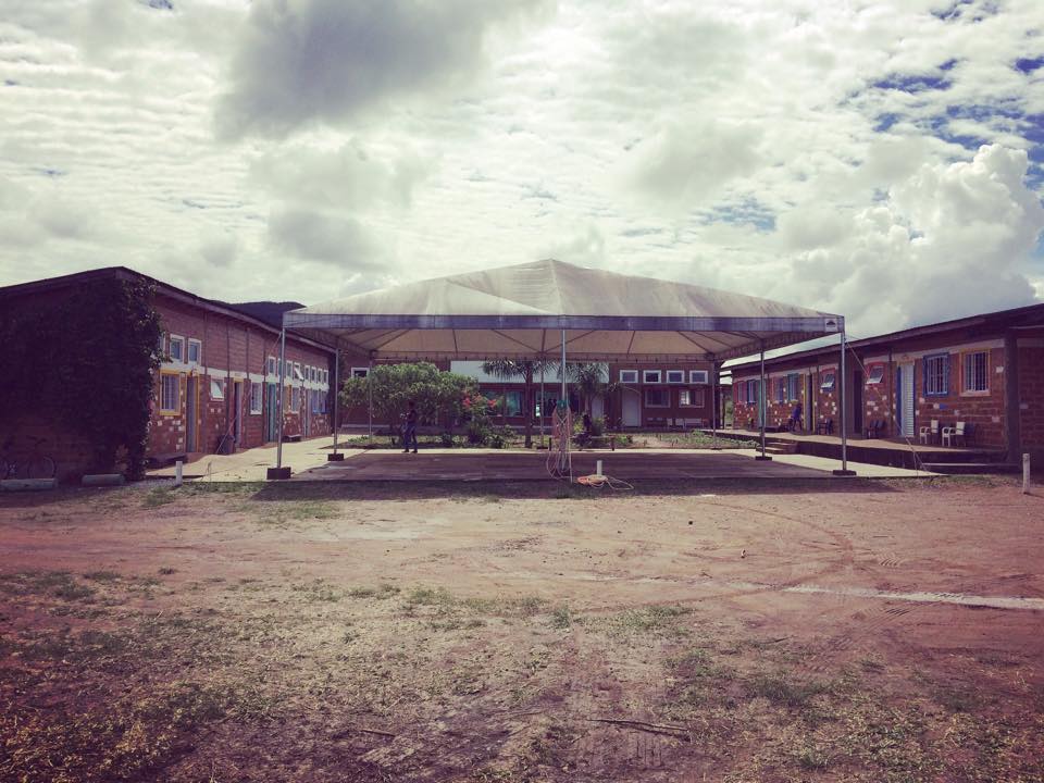 Fachada da escola Vila Verde, em Goiás. Um amplo pátio com vegetação do cerrado em frente a um prédio simples com tijolos à vista e janelas coloridas. No centro da imagem, uma grande tenda.